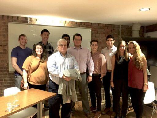 9 Campbell students gathered around Robert Azar, a white-haired man in a light collared shirt. All in front of a whiteboard in a low-ceilinged room.